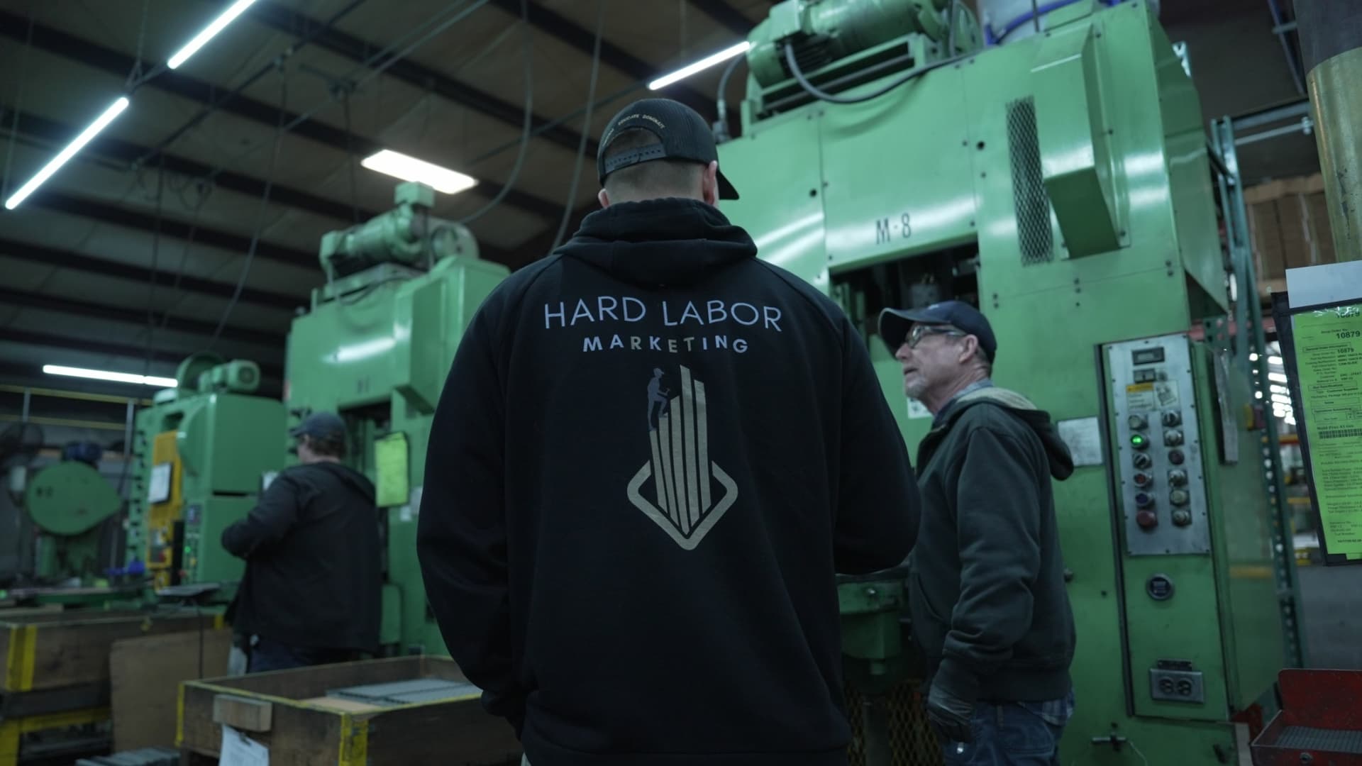 Hard Labor Marketing team member in branded hoodie talking with a press operator beside a green sintering press